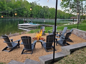 A lakeside seating area with Adirondack chairs, a fire pit, and a dock with a boat.