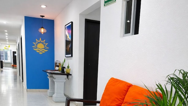 Eine Hotelhalle mit einer Rezeption, einer blauen Wand mit Sonnensymbol, einem weißen Sofa mit orangefarbenen Kissen und einer Zimmerpflanze.