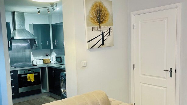 A modern living room with a beige sofa, a kitchen in the background, and a white door. - The Luxe Loft Apartment. (Wolverhampton)