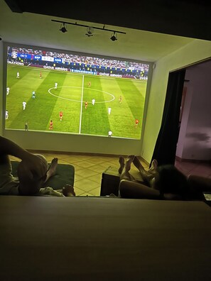 Ein Zimmer mit einem großen Fernseher, der ein Fußballspiel zeigt, einem Bett und einem Fenster mit Blick nach draußen.