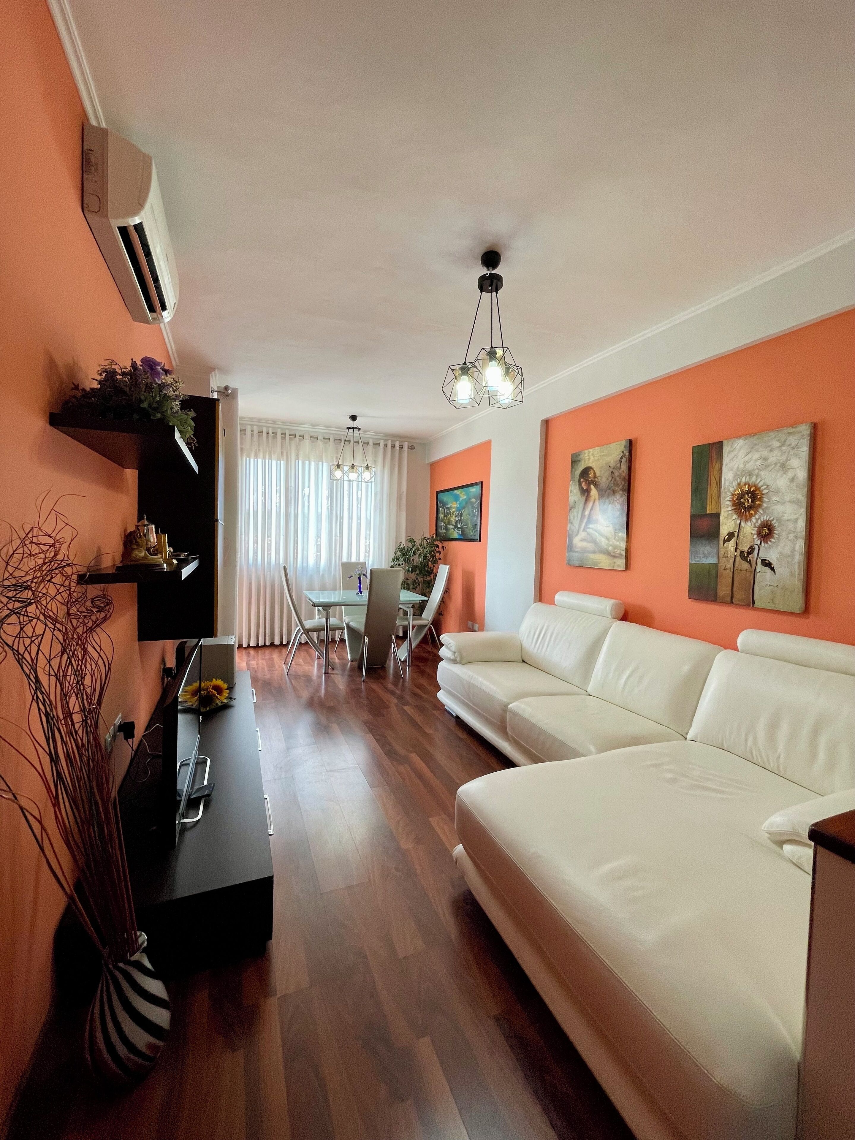 A living room with a white sofa, a black coffee table, and a dining area with a glass table and chairs. There is a ceiling light fixture and artwork on the walls.
