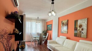 Ein Wohnzimmer mit einem weißen Sofa, einem schwarzen Couchtisch und einem Essbereich mit einem Glastisch und Stühlen. Eine Deckenleuchte und Kunstwerke sind an den Wänden angebracht.