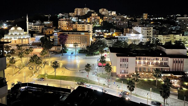 A nighttime cityscape with illuminated buildings, a mosque with a dome and minaret, and a central open plaza with palm trees and a crosswalk. - Illyria Square - City Center Apt - WiFi 500 mb/s (Durrës)