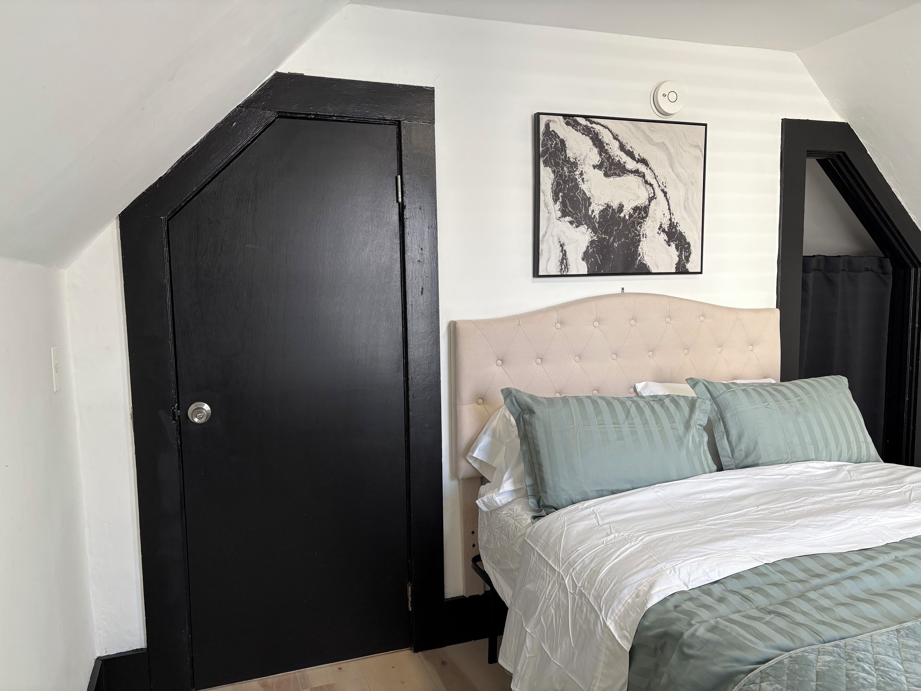 Ein Schlafzimmer mit einer dunklen Tür, einem gesteppten Kopfteil, einem Bett mit weißer und grüner Bettwäsche sowie einem Kunstwerk mit Marmormuster an der Wand.