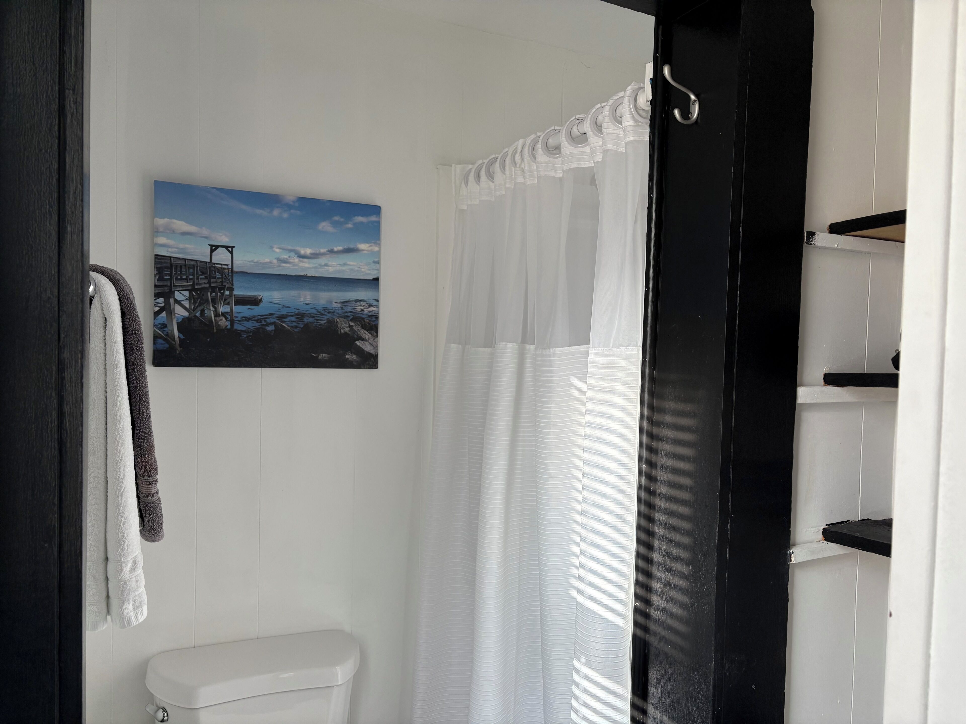 Ein Badezimmer mit Toilette, Handtuchhalter mit Handtüchern, einer weißen Duschvorhang und einem Bild von einem Steg und Wasser an der Wand.