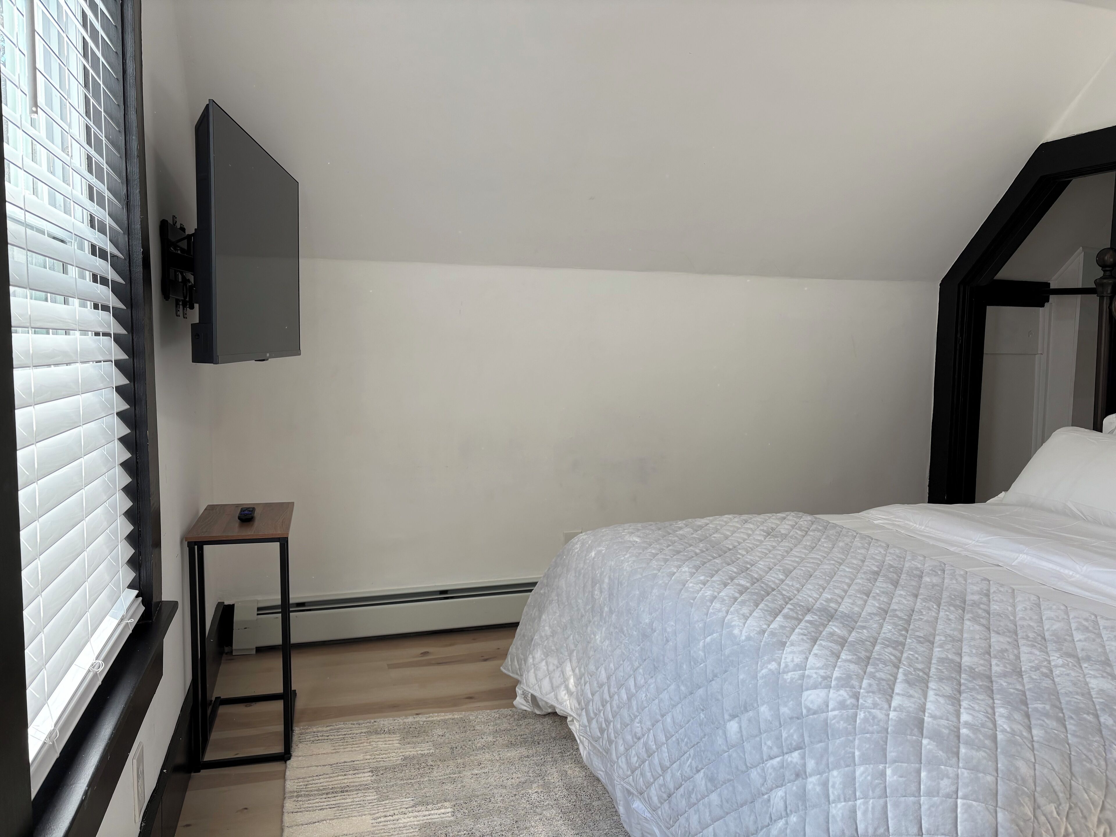 Une chambre à coucher avec un lit, un téléviseur fixé au mur, une petite table et une fenêtre avec des stores.