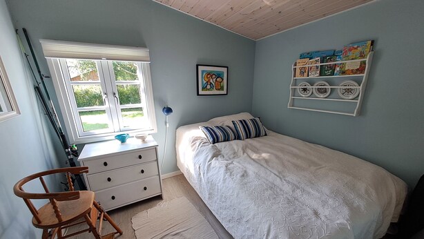 Un dormitorio con cama, cómoda, silla, ventana y estante adosado a la pared con objetos decorativos.