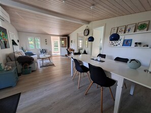 A spacious living area with a dining table, chairs, and a sofa. There is a kitchen visible in the background. - Nature, peace and coziness near forest and beach (Nakskov)