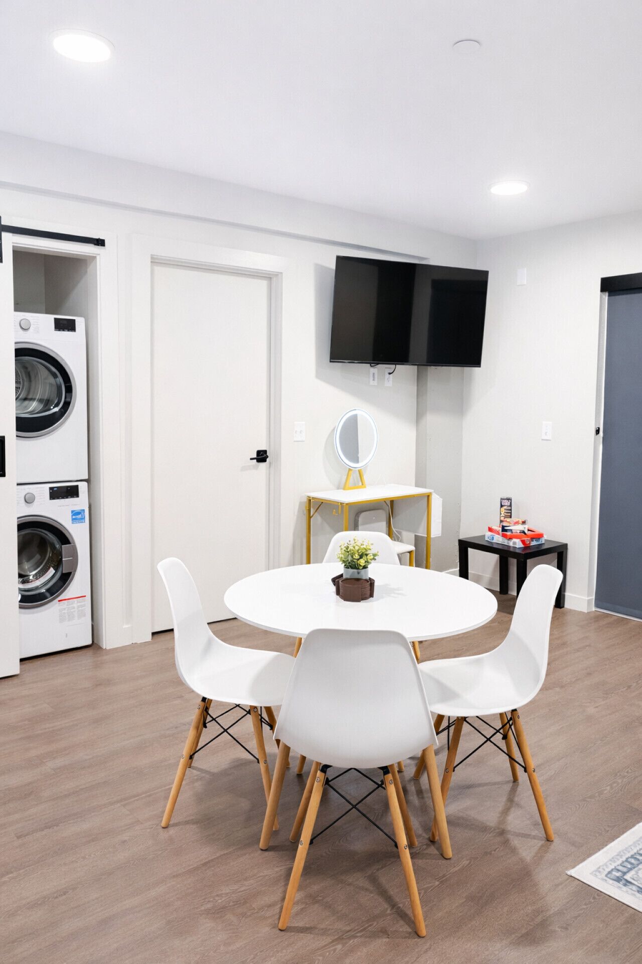 Una habitación moderna con una mesa de comedor redonda, sillas blancas, un televisor de pantalla plana, una lavadora y una pequeña mesa con espejo y jarrón.