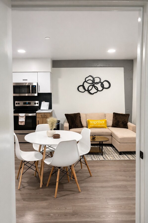 Un espace de vie moderne comprenant un coin repas, un canapé avec des coussins, une œuvre d’art murale et une cuisine en arrière-plan.