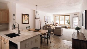 Eine geräumige Küche mit Kochinsel, ein Essbereich mit Tisch und Stühlen, ein Wohnzimmer mit Sofa und Fernseher sowie große Fenster für natürliches Licht.