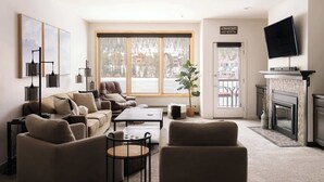 Ein gemütliches Wohnzimmer mit einer Couch, Sesseln, einem Couchtisch, einem Kamin, einem Fernseher und einem großen Fenster mit Blick auf Gebäude.