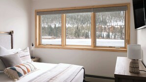 Ein Schlafzimmer mit großem Fenster, einem Bett mit vielen Kissen, einem Nachttisch mit Lampe und einem an der Wand montierten Fernseher.