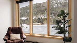 Ein Zimmer mit einem großen Fenster mit Blick auf eine verschneite Landschaft und einen Wald. Ein Ledersessel mit Kissen steht in der Nähe des Fensters.