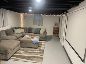 A living room with a sectional sofa, a small ottoman, and a coffee table. There is a white wall with a shelf holding books and a bottle. - Spacious 4-bedroom Sturgis Motorcycle Rally rental with AC located in  Spearfish (Spearfish)