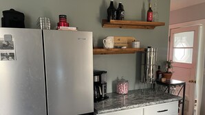 Une cuisine équipée d’un réfrigérateur en acier inoxydable, d’une machine à café, d’une bouilloire et d’une étagère où sont posées des bouteilles et une plante en pot.