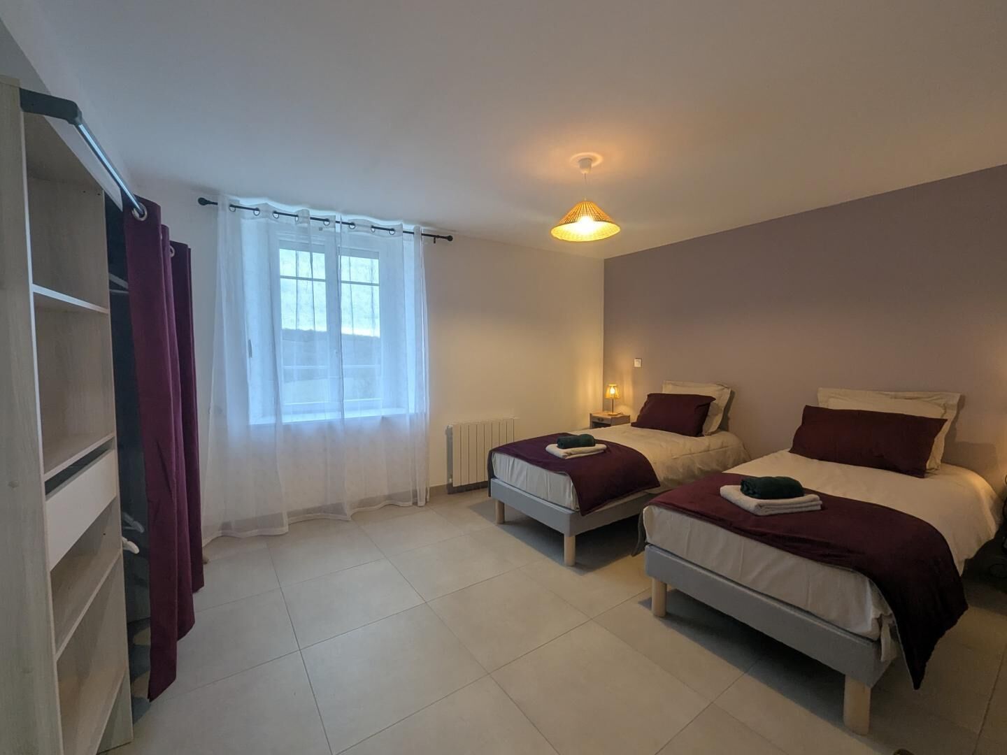 A bedroom with two beds, a wardrobe, a window with sheer curtains, and a ceiling light.