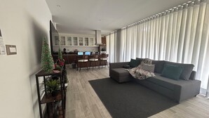 Un salon moderne avec un canapé gris, une table basse et une petite table d’appoint sur laquelle est posée une plante. On aperçoit à l’arrière-plan un coin cuisine avec un ordinateur portable sur le comptoir.