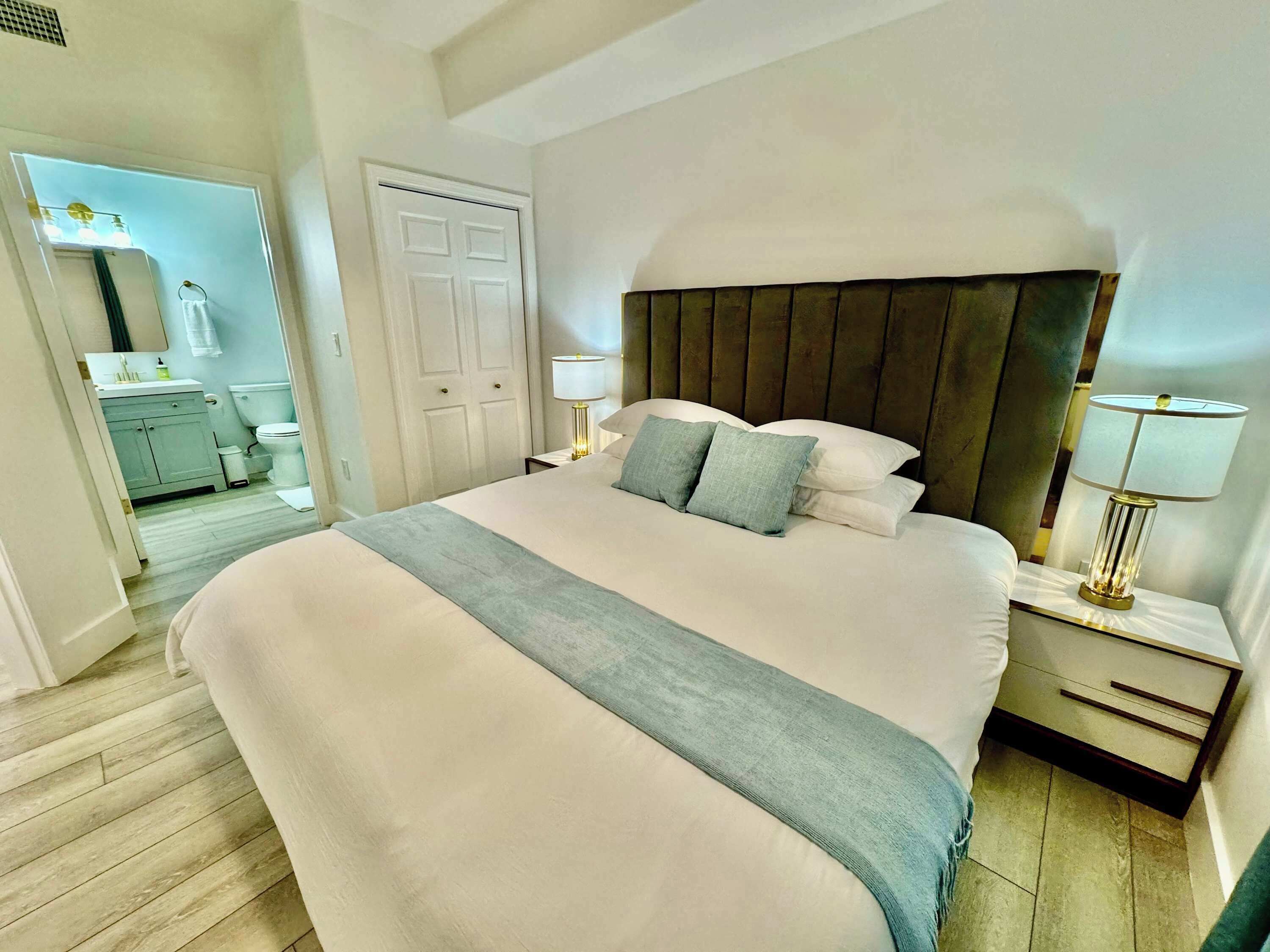Ein Hotelzimmer mit einem großen Bett, einem hölzernen Kopfteil, zwei Nachttischen mit Lampen, einem durch eine offene Tür erreichbaren Badezimmer und einem Holzboden.