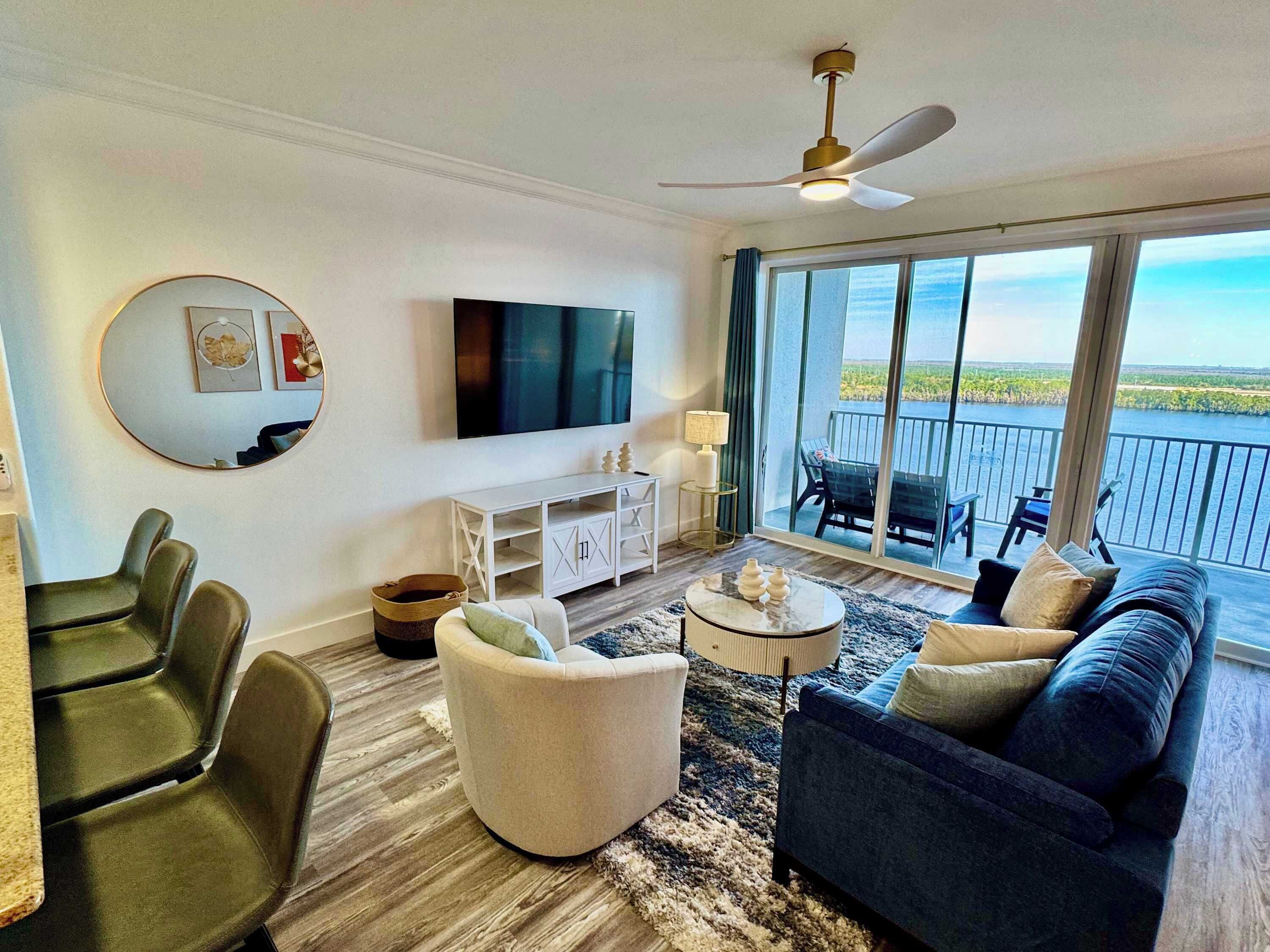 Een moderne woonkamer met een flatscreen televisie, een plafondventilator en een balkon met uitzicht op een waterpartij.