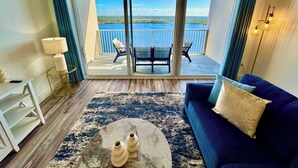 Ein Zimmer mit einem blauen Sofa, einem runden Couchtisch mit zwei Vasen, einem weißen Beistelltisch mit einer Lampe und einem Balkon mit Blick aufs Wasser.