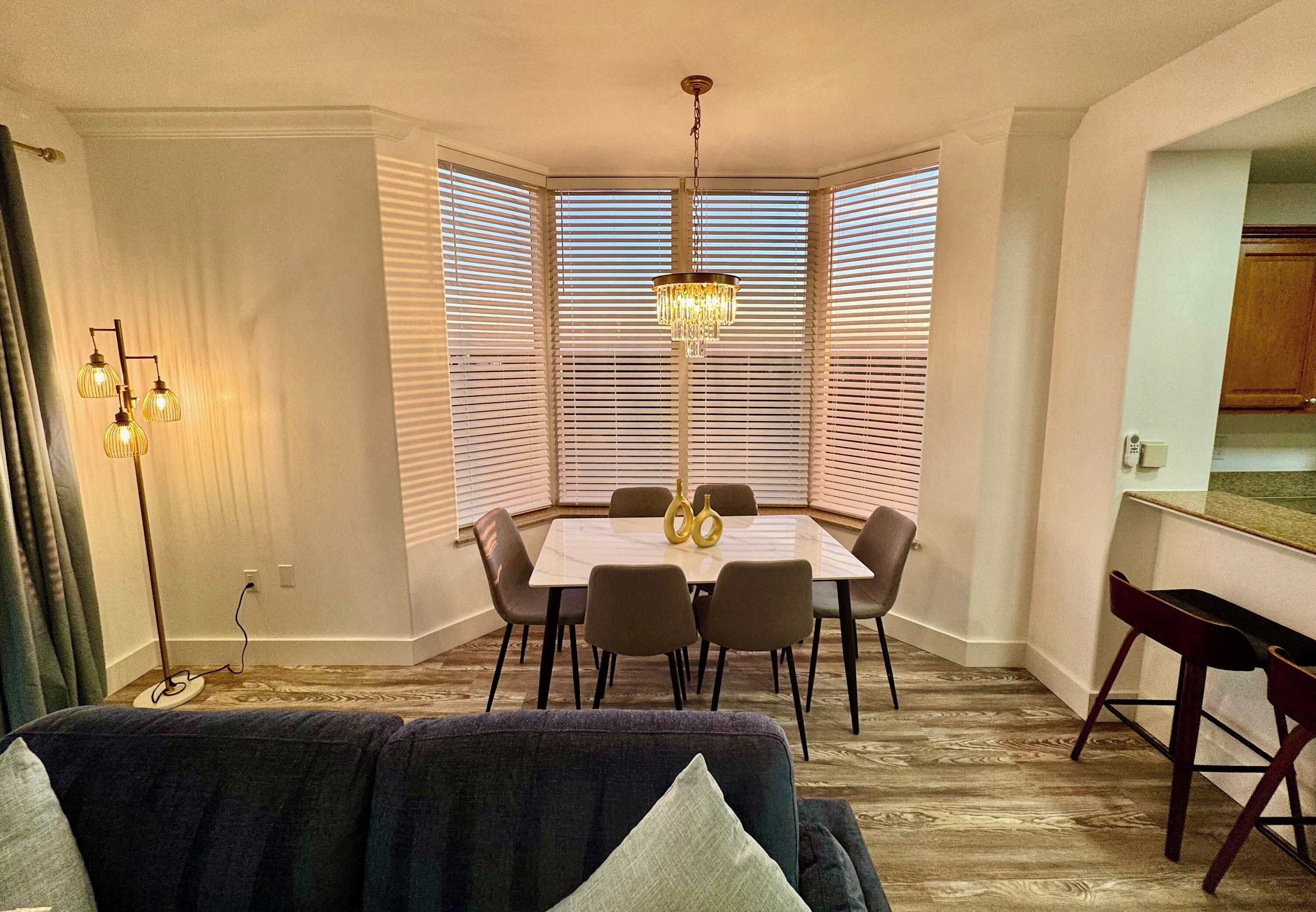 A dining area with a table and chairs, a chandelier, and a view through the window blinds.