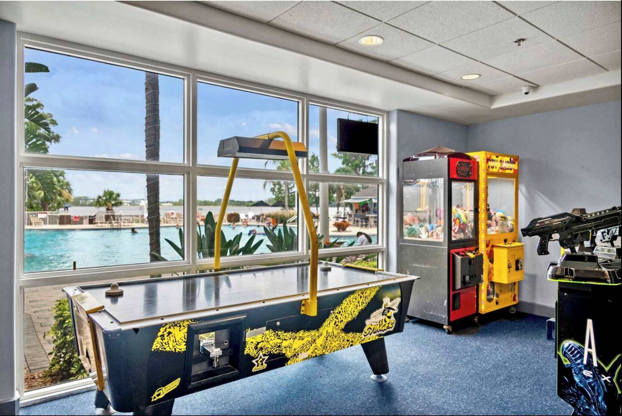 A pool table with a yellow backrest, a claw machine, and a pool view through large windows.