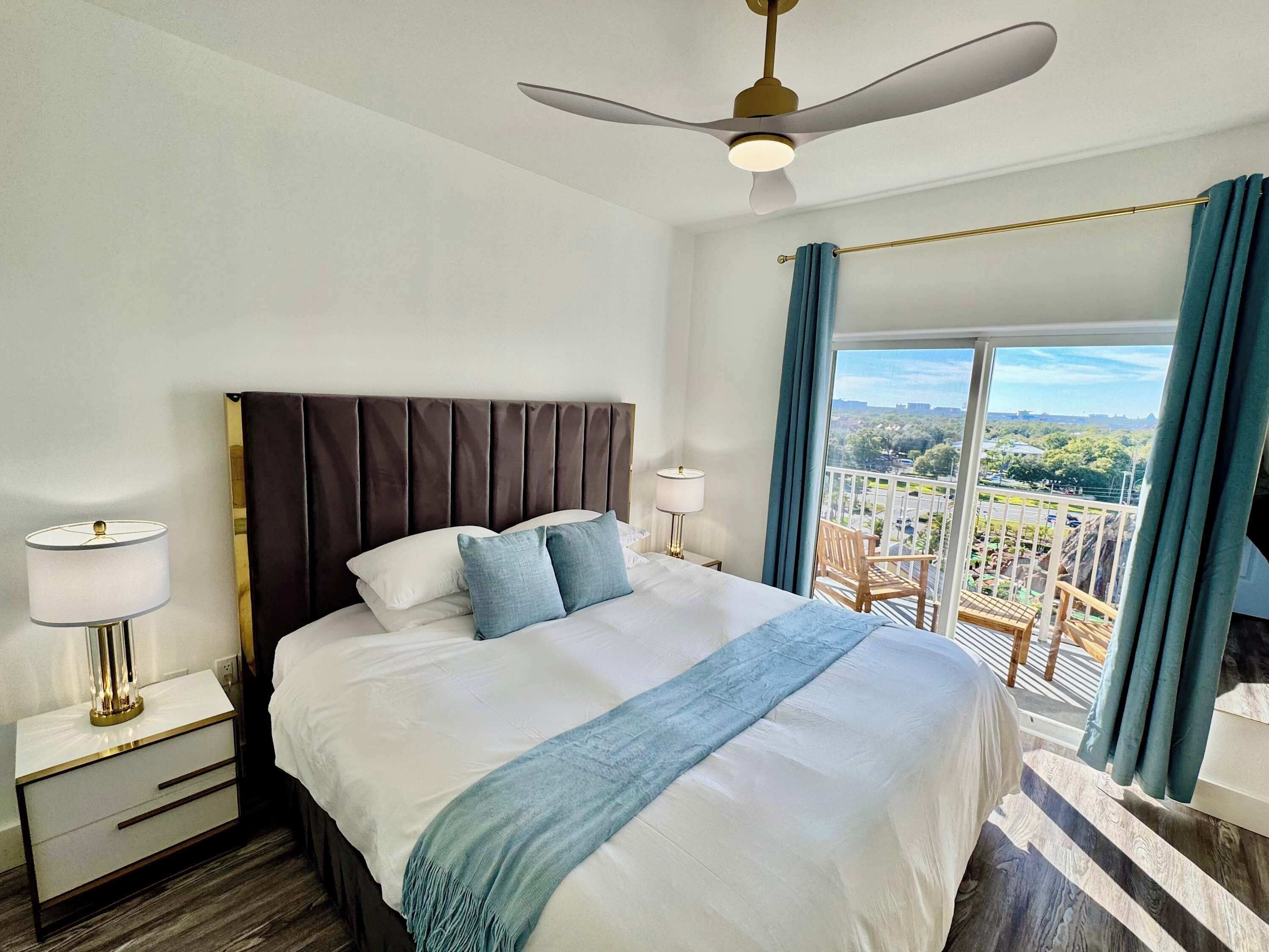 A hotel room with a large bed, a ceiling fan, a nightstand with a lamp, and a balcony with a view of the city.