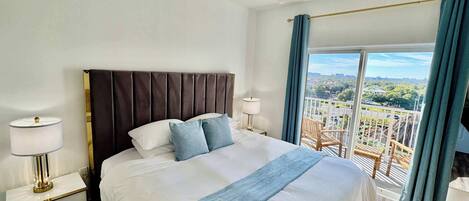 A hotel room with a large bed, a ceiling fan, a nightstand with a lamp, and a balcony with a view of the city.