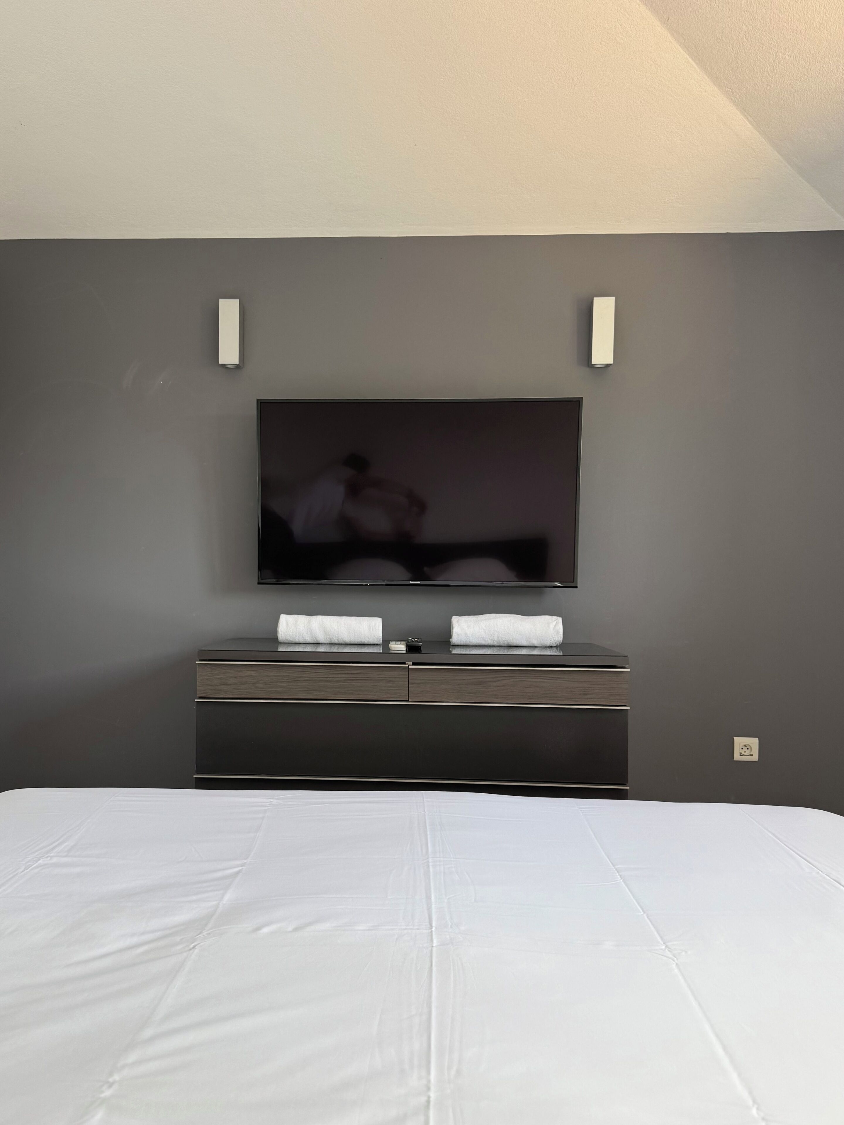 Una habitación de hotel moderna con una cama grande, un televisor de pantalla plana montado en la pared y un aparador de madera oscura con dos toallas.