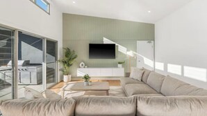 A modern living room with a large sectional sofa, a flat-screen TV mounted on the wall, and a sliding glass door leading to an outdoor area. - Luxe Coastal Escape. Walk to beach, cafe and shops. (Crescent Head)