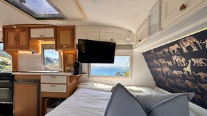 A compact RV interior with a bed, a TV, a kitchenette, and a window with a view of the sea. - Goat-Island Beach Front Get-Away! (Leigh)