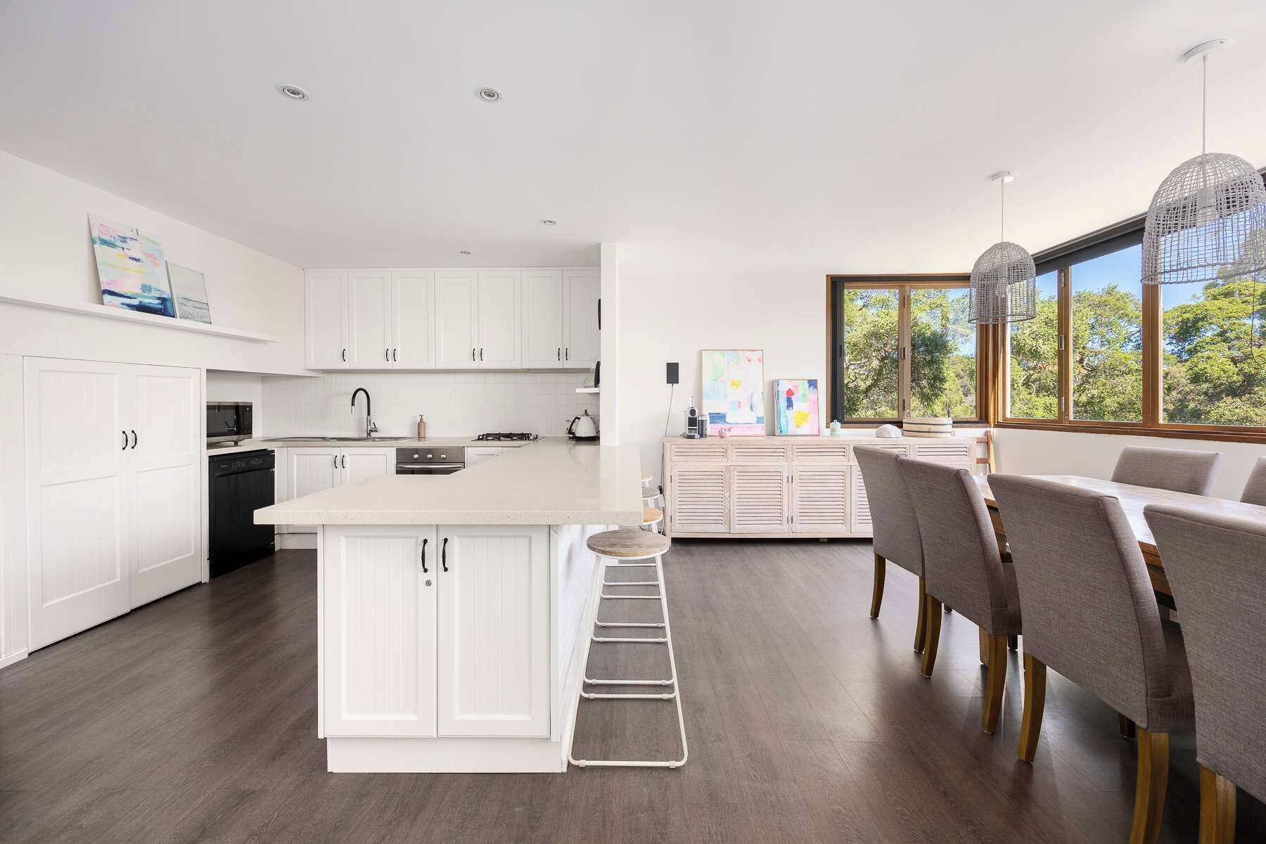 A modern kitchen with white cabinetry, a central island, and a dining area with a view of trees through large windows.