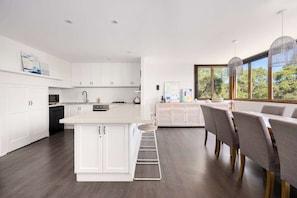 A modern kitchen with white cabinetry, a central island, and a dining area with a view of trees through large windows. - Tuckey Terrace | Heated Pool, Spa & Bay Views (Sorrento)