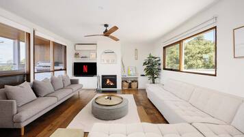 A modern living room with a fireplace, a ceiling fan, and large windows.