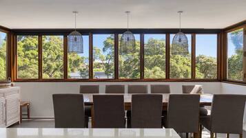 A dining area with a large table and chairs, large windows with a view of trees, and hanging pendant lights.