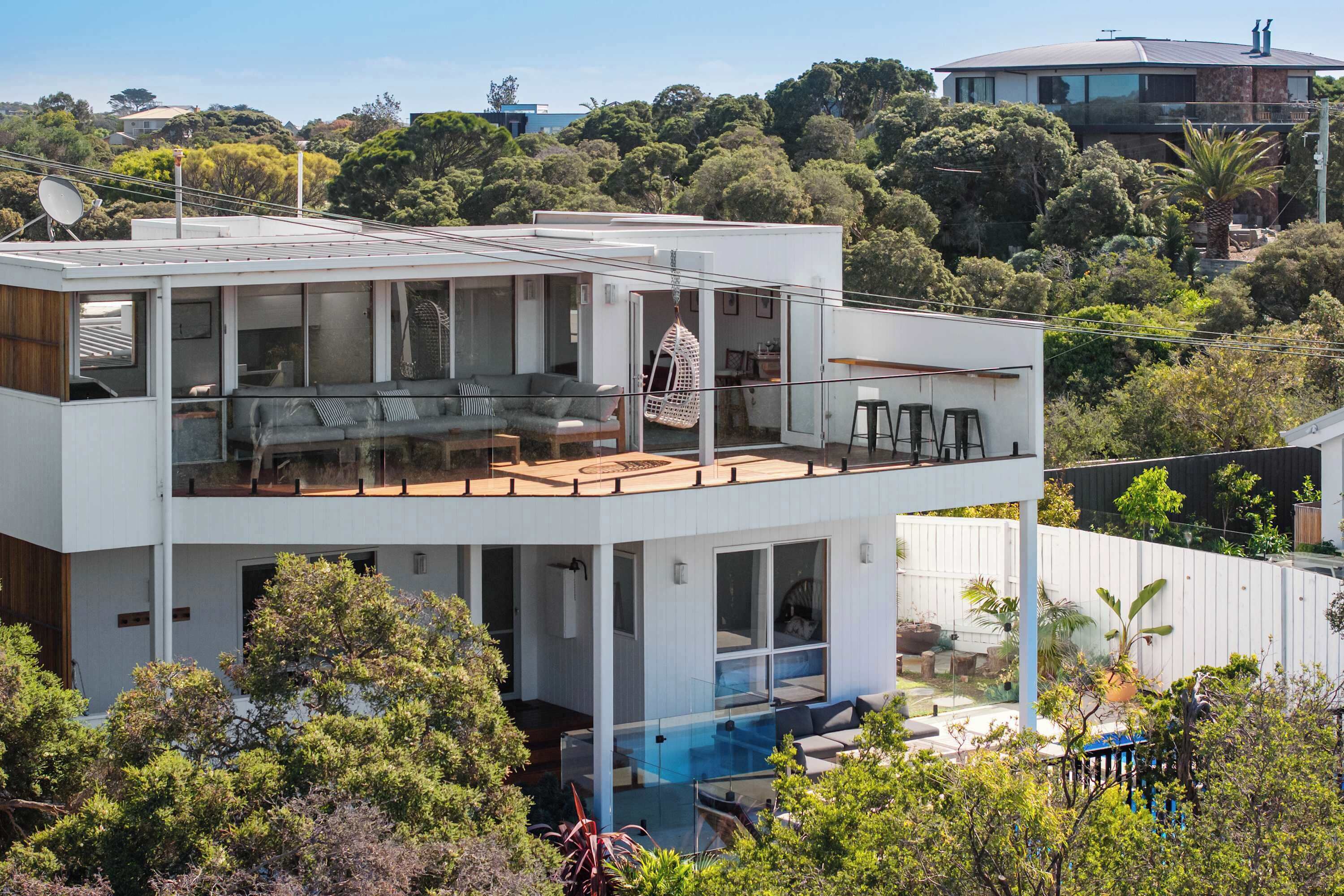 A modern two-story house with a glass balcony, a pool, and lush greenery surrounding it.