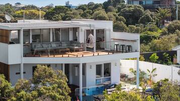 A modern two-story house with a glass balcony, a pool, and lush greenery surrounding it.