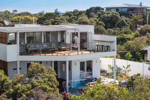 A modern two-story house with a glass balcony, a pool, and lush greenery surrounding it. - Tuckey Terrace | Heated Pool, Spa & Bay Views (Sorrento)