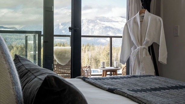 A bedroom with a large window offering a mountain view, a bed with a dark blanket, a white bathrobe hanging on the wall, and a small table with two cups.