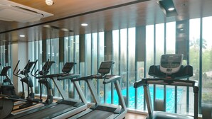 Ein Fitnessstudio mit Laufbändern, Ergometer und Blick auf ein Schwimmbecken durch große Fenster.