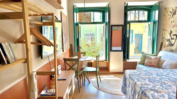 A cozy room with a bed, a desk, and a chair. There are windows with green shutters and a view of the outside. - Rememorar 01 (Salvador)