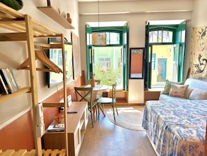A cozy room with a bed, a desk, and a chair. There are windows with green shutters and a view of the outside.
