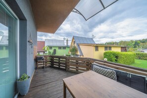 Una terraza de madera con una mesa, bancos, un ventanal y vistas a casas coloridas al fondo.