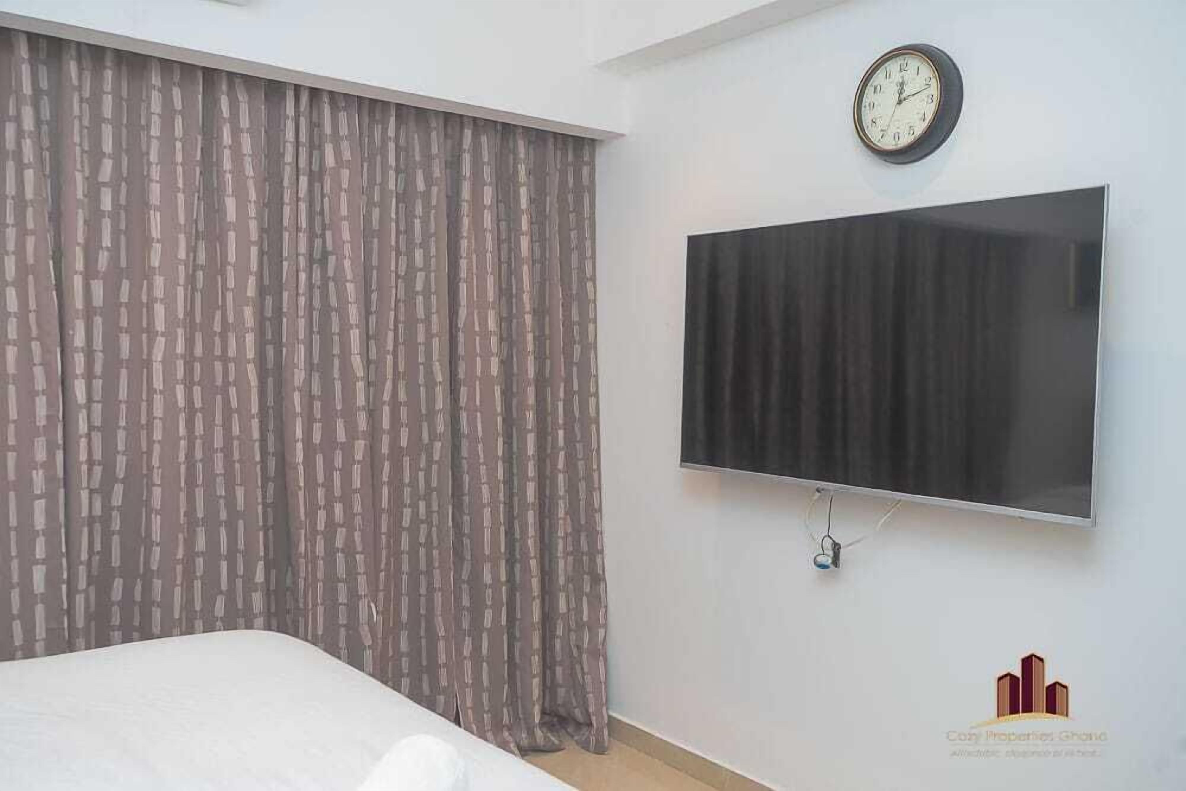 A bedroom with a bed, curtains, a wall-mounted TV, and a clock.
