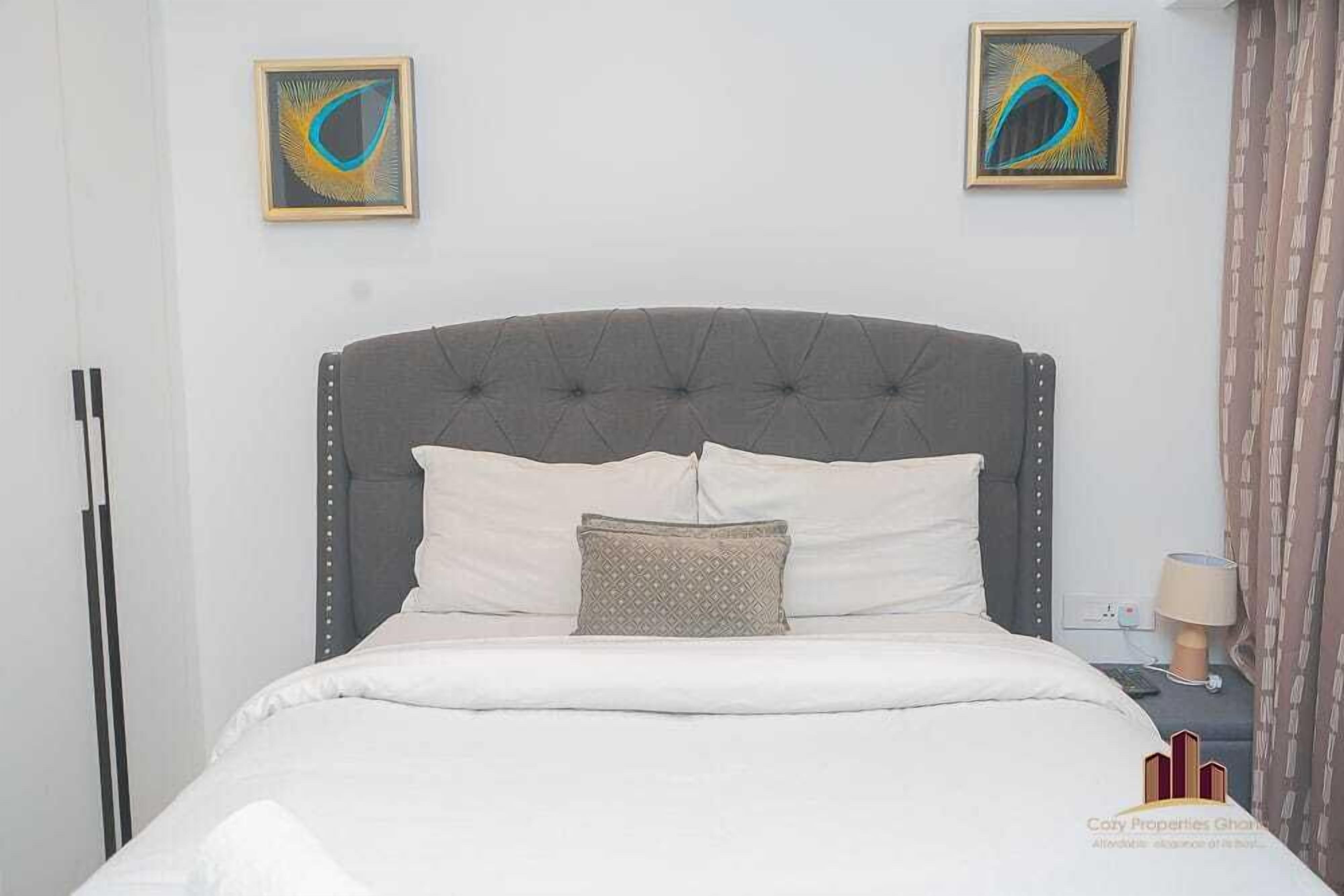 A neatly made bed with a grey upholstered headboard, two pillows, and a decorative pillow. There are two framed artworks on the wall above the bed.