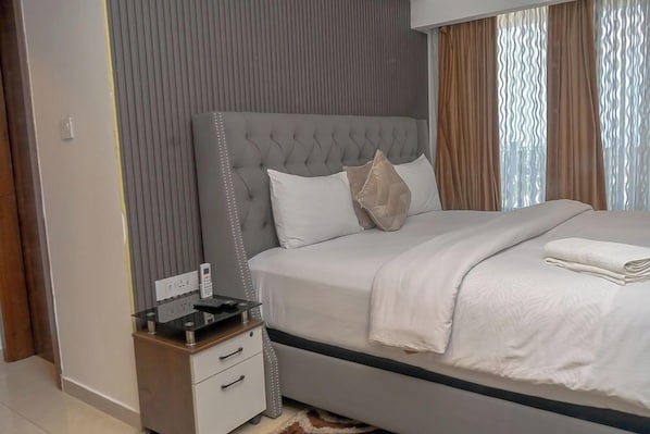 A modern bedroom with a tufted headboard, a bedside table with a phone, and a neatly made bed with white bedding. - Solaris Apartments luxury by Cozy (Accra)