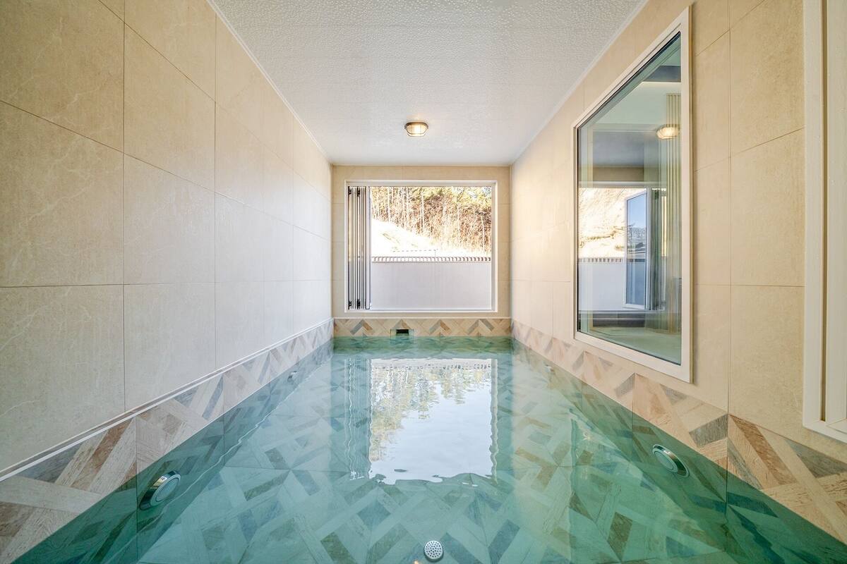Un couloir avec une piscine dont le fond est en verre, des murs recouverts de carreaux beiges et une fenêtre donnant sur des arbres.