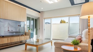 A modern living room with a flat-screen TV, a sofa, a coffee table, and a balcony with a view. - Gyeongju Awesomeway Pool Villa (Gyeongju)