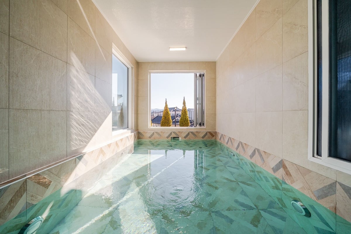Une piscine intérieure étroite, avec une grande fenêtre offrant une vue sur les bâtiments et les arbres.
